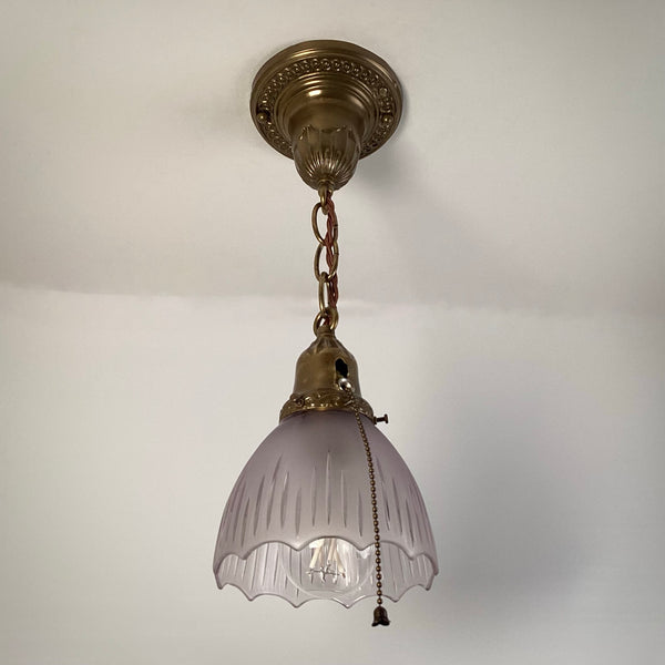 1920s antique brass pendant light with frosted fluted glass shade and pull-chain, hanging from ornate ceiling canopy.