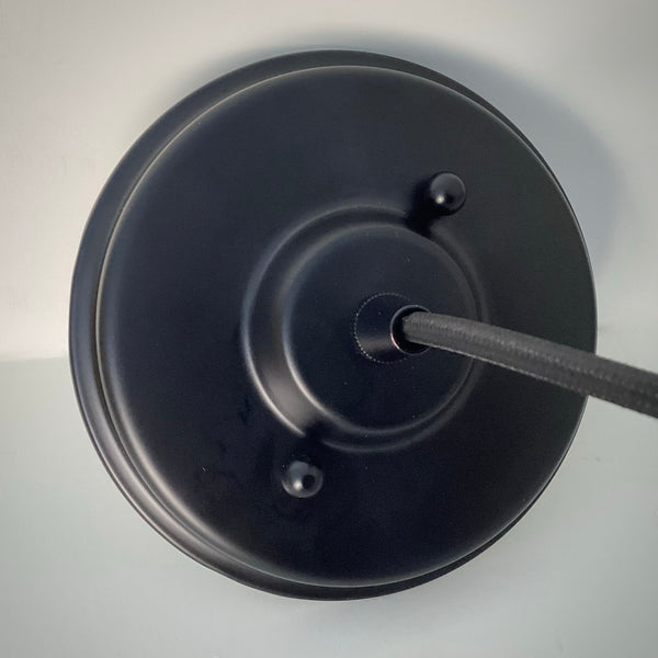A top-down close-up view of a matte black circular ceiling canopy with two decorative mounting nuts and a black fabric-wrapped power cord extending from the center.