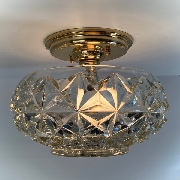 Illuminated mid-century crystal flush mount ceiling light with diamond starburst patterned glass shade and polished brass canopy, showcasing warm reflective glow — Vintporium.
