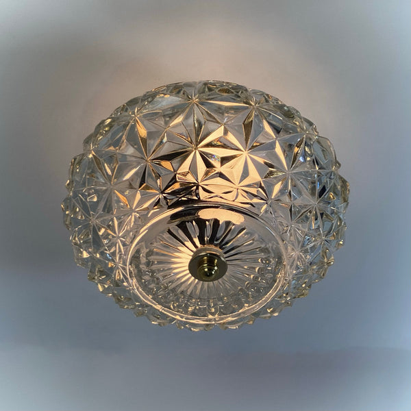 View from below of an illuminated mid-century crystal flush mount ceiling light, showing the detailed starburst and diamond-cut glass pattern with brass finial centerpiece — Vintporium.