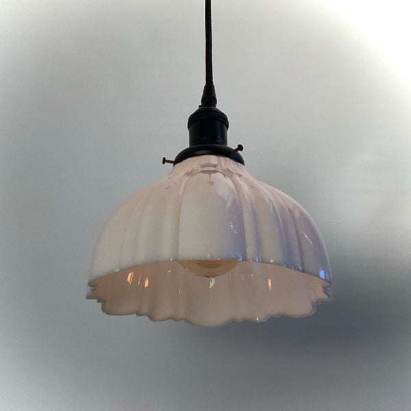 A fully assembled pendant light featuring a vintage-style white scalloped glass shade held by a black fitter, hanging from a black cord against a neutral wall.
