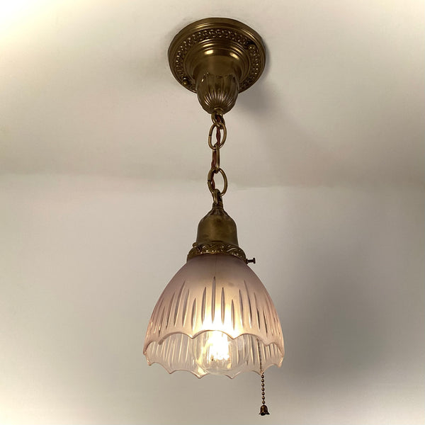 1920s antique brass pendant light with illuminated frosted fluted glass shade and pull-chain, hanging from ornate ceiling canopy.