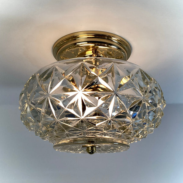 Illuminated mid-century crystal flush mount ceiling light with faceted diamond starburst glass shade and polished brass canopy, casting a warm golden glow — Vintporium.