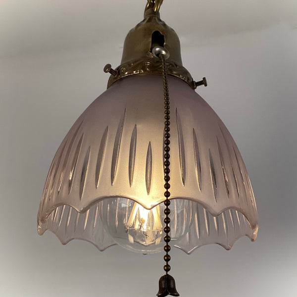 Close-up of a 1920s antique pendant light showing the frosted fluted glass shade, glowing bulb, and brass pull-chain.